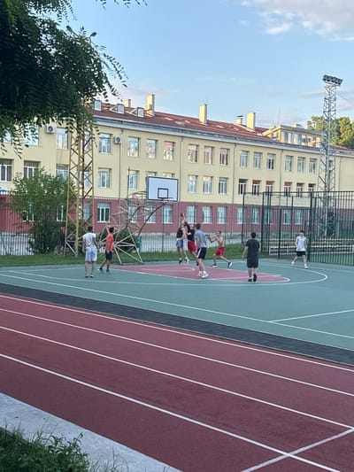 The image depicts an outdoor basketball court located within the city of Shumen, Bulgaria, likely adjacent to a school facility, as suggested by the building in the background. The court surface is a vibrant green with red accents around the perimeter and a red central area. Several individuals, appearing to be young men, are engaged in a basketball game. One person is mid-air, attempting a shot or rebound, while others are positioned around the court, actively defending or preparing to receive a pass. The individuals are dressed in athletic wear, including shorts and t-shirts. The surrounding environment includes a metal fence, a treeline, and a tall metal structure with lights, possibly for evening use of the court. In the foreground, a running track with red surfacing and white lane markings is visible. The sky is partly cloudy and appears to be late afternoon or early evening.