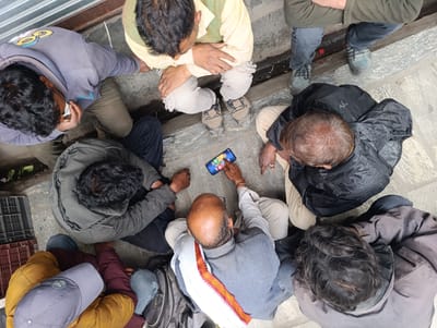 A group of six individuals are gathered around a smartphone laid flat on the ground. The people are seated on a stone-tiled surface, with a metal grate and a weathered wooden structure in the background, suggesting an outdoor setting, possibly in a marketplace or public walkway. The time of day appears to be daytime, with ample natural light. The weather is not explicitly discernible, but the attire suggests it could be cool.
One person, wearing a light yellow kurta and green vest, is leaning over the phone, seemingly demonstrating something or playing a game. Another person, in a grey hoodie and dark pants, is also looking at the phone, with a tissue held to their nose. A third person in a black puffer jacket and beige pants is partially visible. The remaining individuals are seated around them, their faces mostly obscured from this overhead perspective. One person is wearing a yellow jacket and a grey baseball cap. Another person is wearing a grey jacket. The final person is wearing a dark grey jacket and is looking down.
The smartphone screen displays a colorful game interface, with icons that resemble dice and game boards. The overall mood appears casual and engaging, with the group focused on the shared activity on the phone. The location context of Manali, India, is provided, hinting at a potentially tourist-frequented area given the stone paving and diverse attire. No discernible text is visible on the clothing or immediate surroundings, other than the icons on the phone screen.