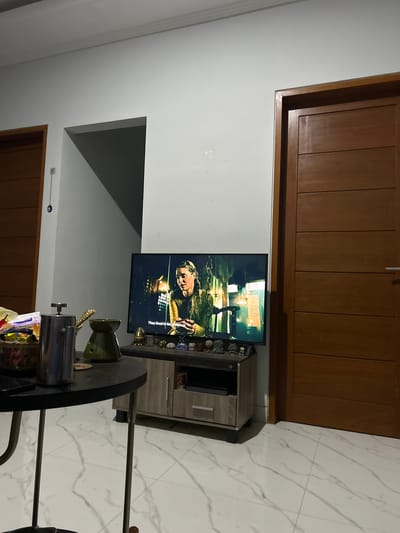 The image captures a home entertainment setup in what appears to be a living room. A flat-screen television is mounted on a wall, displaying a scene with a person dressed in historical attire. The TV is placed above a small, light-wood entertainment cabinet. On top of the cabinet, a collection of decorative items, possibly figurines or small sculptures, are arranged. To the right of the television, white tiled flooring with subtle grey veining is visible.
In the foreground, a dark, round table is partially visible, with a metallic coffee pot or dispenser and some packaged snacks on it. The room's walls are painted white, and a large wooden door with vertical paneling is seen on the left side of the image. Another similar wooden door is partially visible at the bottom. The overall lighting suggests it is daytime. The scene appears calm and domestic, typical of a residential space. There is some text on the TV screen, "They lived in his presence." The location context provided is Sukawati, Indonesia.