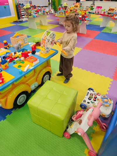 A child in a beige long-sleeve shirt and brown pants stands in a children’s play area. The floor is covered in colorful interlocking foam tiles. To the left of the child is a blue table with a yellow base shaped like a car, covered with Lego blocks and other toys. A large, green, cubic seat is positioned next to the car-shaped table. To the right, there are two infant walkers. Additional play structures and toys are visible in the background, including a toy city. “МЕРКУРИИ” is visible on a box in the bottom left corner. The city is Khimki, Russia.