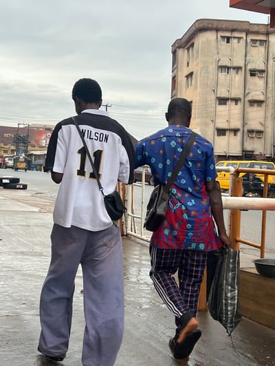 The image shows two men standing on a wet, possibly concrete, surface. The man in the foreground, closer to the camera, is wearing a white and black long-sleeved shirt with "WILSON" and the number "11" printed on the back in black and gold. He has a black crossbody bag. He is wearing grey trousers. The second man is further away, wearing a blue and red patterned short-sleeved shirt and checkered trousers with white stripes down the side. He also has a black crossbody bag. Behind the men, there is a building with multiple windows, a yellow bus, and some metal railings. The scene appears to be outdoors, possibly in an urban or semi-urban area, given the building and the bus. The ground is wet, suggesting it might have recently rained or there's a water source nearby. The sky is overcast, indicating a cloudy day. There is text visible on the shirt of the man in the foreground: "WILSON 11". There are also some signs of shops and advertising in the background, some with text that is not clearly legible. The overall mood seems neutral, with the men appearing to be in transit or waiting. The context provided places this scene in Nkpor, Nigeria.