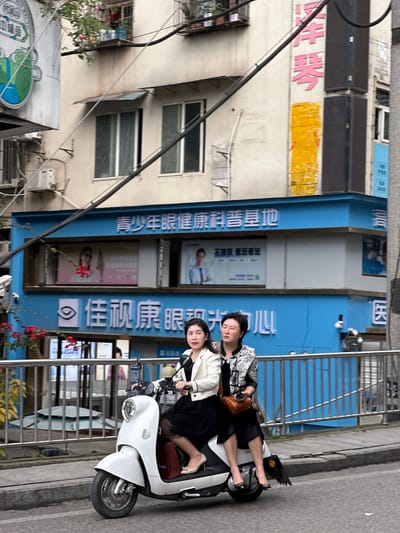 Two women on a white scooter sit on a street in Jiangjin, China. The woman in front wears a white jacket and the woman in the back wears a patterned top. They are paused in front of a building that is light-colored with blue trim and writing, including the words "青少年眼健康科普基地". There is a steel barrier in front of the building. A variety of signs are on the building. Street foliage grows around the barrier. A utility pole with wiring is visible on the left.