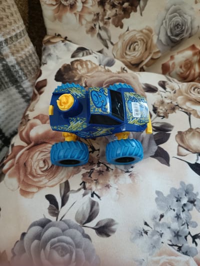A blue toy monster truck with yellow accents and large blue tires sits on a floral-patterned surface. A tag on the side of the truck reads "TORNADO". The background consists of a patterned fabric that appears to be upholstery. There is no information within the image to suggest a location in Băcioi, Moldova.