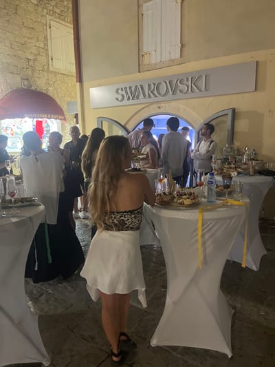 The image depicts a social gathering outside a Swarovski store in Budva, Montenegro. Several individuals are observed mingling, with some standing near tables laden with food and drinks, while others are engaged in conversation or observing the surroundings. A sign above the store entrance clearly reads "SWAROVSKI". White tablecloth-covered tables are arranged, displaying platters of appetizers, glasses, and bottles of beverages, including a prominent bottle of water. The ground appears to be paved with cobblestones, suggesting an outdoor urban setting. The overall atmosphere suggests an event or reception held in the evening.