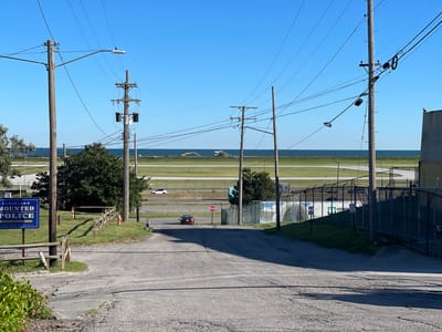 The image depicts a view looking down an asphalt road toward a body of water under a clear blue sky. On the left side of the road, a blue sign reads "CLEVELAND MOUNTED POLICE UNIT." A wooden fence runs along the grassy area beside the sign. Further down the road, a red car is stopped at a stop sign. Beyond the road, a fenced area is visible, and in the distance, a flat expanse of green land meets the water, with two yellow excavators working in the distance. Utility poles and wires are prominent throughout the scene, crossing overhead. On the right side of the image, a large industrial-looking building or structure is partially visible.