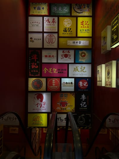 This media file showcases a vibrant display of food stalls, likely in a marketplace or hawker center, given the numerous signs advertising various Asian dishes. The scene is illuminated by bright, overhead lighting, suggesting an indoor or evening setting. The overall aesthetic is rich with color and text, with many signs featuring Chinese characters and English translations, hinting at a multicultural environment.
The subjects are primarily the food stalls themselves, each adorned with eye-catching signage. These signs are rich with visible text, detailing the names of the dishes and establishments, such as "POPORCO MACAU PORK BUN," "KONG TAI SINGAPORE FAMOUS HOKKIEN PRAWN MEE," "KIN KIN CHILI PAN MEE," "SENG KEE CHICKEN RICE," and "HO WENG KEE BEEF NOODLE." There are also signs that appear to be for drinks, like "LAYUMCHA." The variety of food advertised suggests a focus on Chinese and Southeast Asian cuisine.
While no people are clearly visible in the foreground, the presence of escalators in the lower portion of the image suggests a bustling public space, possibly a shopping mall or a large indoor market. The location context of Kuala Lumpur, Malaysia, further reinforces the idea of a vibrant urban setting known for its diverse street food culture. The overall impression is one of an energetic and appetizing culinary destination.