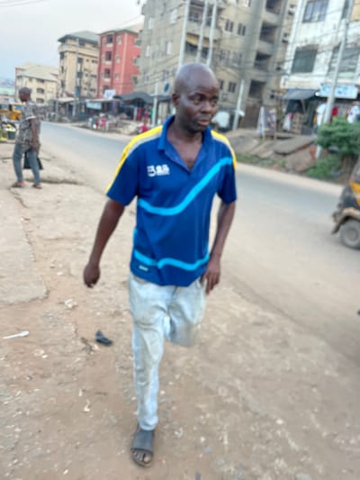 An outdoor, daytime photograph captures a man standing on an unpaved, dusty path next to a road, with buildings and another person in the background. The image is taken from a slightly low angle and is rotated counter-clockwise.
The primary subject is a bald, dark-skinned man of apparently middle age or older, looking towards the viewer's left side. He wears a blue short-sleeved polo shirt with distinctive light blue and yellow stripe designs across the chest and shoulders. On the left side of his chest, a white logo or text that appears to be "3B" is visible. He is also wearing light-colored pants, which appear somewhat soiled or worn, and dark sandals or flip-flops on his feet. His expression is neutral or slightly serious. His left arm is bent at the elbow, while his right arm is mostly obscured but appears to be by his side.
The ground where he stands is a mixture of dirt and gravel, with visible debris including a white plastic spoon and a dark, unidentified object that might be a discarded slipper or piece of trash. To his right and behind him, a section of a paved or hard-packed dirt road is visible. Further down this road, another person can be seen walking, smaller in the frame, wearing a patterned shirt and dark pants.
In the background, on the left side of the frame, a cluster of multi-story buildings stands, suggesting an urban or semi-urban environment. Some buildings are light-colored (grey or off-white), while one in the mid-ground is a reddish-brown hue. Visible electrical wires or pipes run along the exterior of some structures. Small commercial establishments or signs are faintly discernible near the buildings. A part of a yellow vehicle, possibly a tricycle or rickshaw, is visible at the far upper right edge of the frame. The sky is light, indicating daylight conditions.