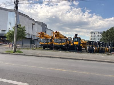 A fleet of yellow construction vehicles, including several cranes and forklifts, are parked in an outdoor lot adjacent to a street in Tbilisi, Georgia. The vehicles are arranged in rows behind a black chain-link fence. In the background, a multi-story white building with a modern design is visible, as is a taller residential building with balconies. The sky is partly cloudy. A utility pole with numerous wires stands on the left side of the frame, near a small, green tree. The street in the foreground is asphalt with yellow and white markings.
