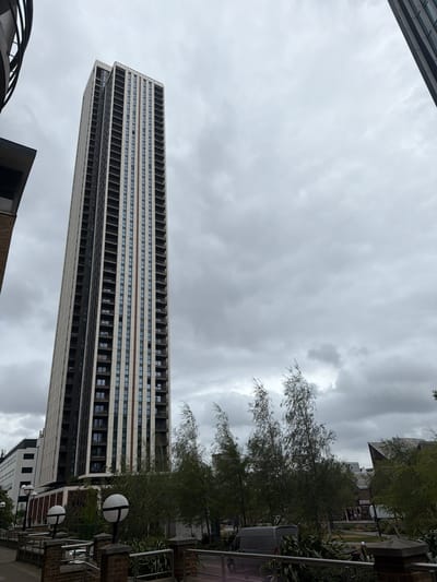 A tall, rectangular building dominates the frame against a cloudy sky. The building has a repetitive pattern of horizontal lines and window openings. Adjacent to the building are trees and greenery, along with street lamps and elements of urban infrastructure. The scene is set in an urban environment, possibly the city of London, United Kingdom. The details of the architecture and surrounding environment suggest a modern cityscape.