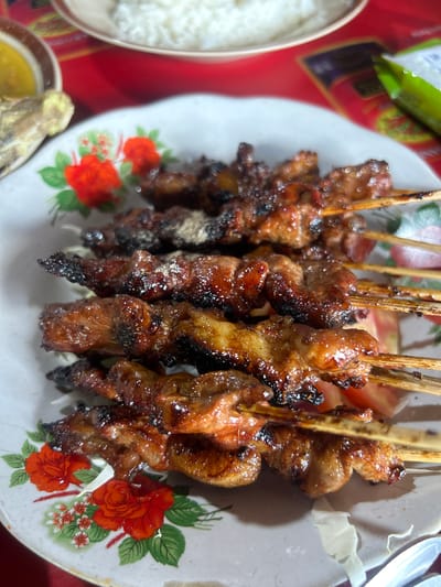 This is a close-up, overhead shot of a plate of grilled meat skewers, likely satay, on a white plate adorned with a floral pattern of red roses and green leaves. The skewers are arranged horizontally on the plate and appear to be made of chunks of seasoned meat, some of which are slightly charred from grilling. The meat has a glossy, well-cooked appearance, with visible grill marks on some pieces. A few pieces of sliced tomato and shredded cabbage are visible underneath the skewers. In the background, out of focus, a bowl of white rice and a small dish containing a yellow liquid or sauce can be seen, along with a red surface that could be a tablecloth. The overall presentation suggests a casual dining setting, possibly a local eatery in Surakarta, Indonesia, known for its satay dishes.