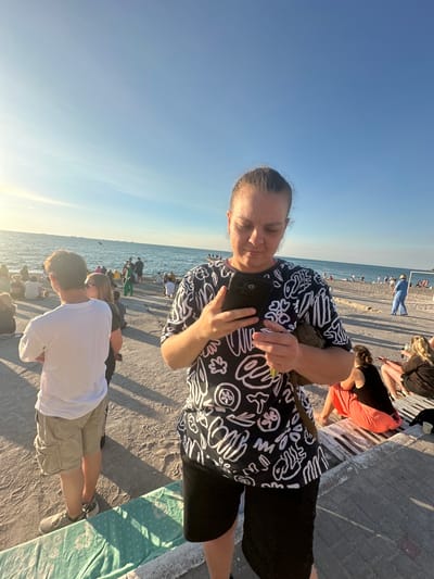 A woman in a black and white patterned shirt and black shorts stands on a concrete promenade, looking down at her smartphone, which she holds in both hands. She is in the foreground, with a beach and the sea visible behind her. People are gathered on the beach and along the promenade, some sitting on benches. The sun is shining, casting long shadows. The location is the Odesa seashore in Ukraine.
