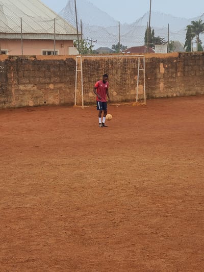 The image shows a person in a red shirt and dark shorts playing soccer on a dirt field. They are wearing white socks and cleats. A soccer ball is near their feet. In the background, there is a soccer goal with a net. Behind the goal, there is a brick wall, and beyond that, some buildings and trees. A fence surrounds part of the field. The sky is overcast, suggesting it might be early morning or late afternoon, or a cloudy day. The location is identified as Barakin Lerawi, Nigeria. There is some text visible on the person's shorts, which appears to say "NAHA".