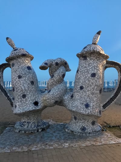 The image shows a mosaic sculpture on a beach, likely in Varna, Bulgaria, given the context. The sculpture resembles two large teapots connected by a serpentine element, all covered in a mosaic of white, grey, and blue tiles. The teapots have handles and spouts. In the background, a sandy beach is visible with rows of white beach umbrellas and lounge chairs, indicating a public beach area. The sea is present beyond the beach, and the sky is a clear, pale blue, suggesting it is either early morning or late evening. There are no people visible in the image.