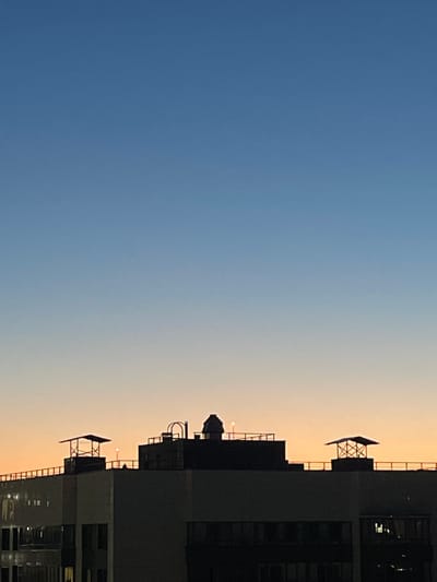 The image depicts a silhouetted view of a building's rooftop against a gradient sky, transitioning from deep blue at the top to a pale orange near the horizon. The rooftop features several structures, including what appear to be chimney caps and a more elaborate, framed canopy with a sloped roof. A faint light source is visible in the distance above the rooftops. The building itself is largely dark, with some illuminated windows visible on the lower levels. The scene is captured from a perspective looking upwards, suggesting the photo was taken from a lower vantage point or another building. There are no people or discernible actions within the frame. The image is likely taken during dusk or dawn, given the sky colors. The location specified, Sinyavino, Russia, provides a geographical context to the urban rooftop scene.