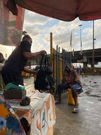 The scene depicts an outdoor commercial area in Lekki, Nigeria, under a partly cloudy sky with a visible low sun. The ground is paved with interlocking bricks and appears wet from recent precipitation. A tattered red canvas canopy partially covers the foreground.

In the left mid-ground, a dark-complexioned male, wearing a black t-shirt and black shorts, stands next to a makeshift stall covered with a faded banner. He holds up a black backpack, from which two dark, stylized plush figures are suspended by their straps. A light blue-green point-of-sale (POS) terminal rests on a grey concrete block on the stall. Earbuds are visible in the male's left ear.

To the right, a second dark-complexioned male is seated on a yellow cubic object. He wears a colorful, patterned short-sleeved shirt, dark cropped trousers, and white and blue flip-flops. He is smiling and looking towards the viewer. A beaded necklace is visible around his neck.

The background features a row of yellow vertical metal barricades. Behind these, a wet road shows a yellow auto-rickshaw (keke napep) in the distance. Further back, an elevated concrete road structure spans the frame, with traffic light poles and multiple flags, including a green, white, and red banner, also visible.