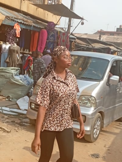 The image displays an outdoor market scene in Fegge, Nigeria, under daylight. A young woman of medium-dark complexion is the central figure, walking from left to right across the foreground. She wears a leopard print headscarf, round spectacles, a short-sleeved collared shirt patterned with brown, white, and black florals, and black leggings. A small dark bag is carried over her left shoulder. Her gaze is directed straight ahead, slightly towards her right.
In the background, to the left of the woman, a market stall exhibits various clothing items. Two mannequins are visible, one displaying a light-colored top, the other a dark olive-green dress. Numerous additional garments, including bright orange and pink trousers, patterned fabrics, and sequined tops, are hung from racks beneath a dark awning. Several individuals, including a man in a plaid shirt and another in a red and blue shirt, are present near the stall. Large, light-colored sacks are piled on the ground next to the market display.
To the woman's right, a silver compact van is partially visible, facing left, with its front headlight and bumper discernible. A wooden utility pole with attached wires stands in the mid-ground. Further in the background, other structures with corrugated metal roofs and light-colored walls are visible under a hazy sky. The ground is unpaved, appearing as dusty, brownish-orange earth.