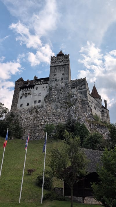 The image depicts Bran Castle, also known as Dracula's Castle, situated on a rocky outcrop in the city of Predeluț, Romania. The castle is a large, stone structure with multiple towers and red-tiled roofs, appearing to be perched atop a steep, rocky hill. Several flags are visible on tall poles in the foreground, set against a grassy slope leading up to the castle. The sky above is partly cloudy with patches of blue. A small, dark-colored building with a wooden roof is partially visible on the right side of the frame, at the base of the hill. The overall scene is a scenic view of the historic castle and its surrounding landscape under a partly cloudy sky.