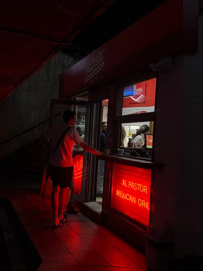 This nighttime image captures a vibrant scene outside and inside a Mexican restaurant in Sofia, Bulgaria, bathed in striking red neon light. The perspective is from an outdoor vantage point, looking towards the brightly lit establishment.

In the foreground, a young man stands, partially obscured from the waist down, dressed in a white t-shirt, dark shorts, and white athletic socks with black stripes. A dark strap from a bag crosses his shoulder, and he stands near a yellow delivery bag with "GI" or "GIO" visible on its side. The ground beneath him is a red-tiled surface, reflecting the neon glow.

The main subject of the image is the restaurant itself. Through its large windows, several individuals are visible inside. One person with dark hair, wearing a white top, stands behind a counter, appearing to be working at a point-of-sale system with a monitor displaying text, possibly "MENU". Another person is partially seen deeper within the establishment. The interior walls are white, possibly brick.

The most prominent feature is the bright red neon sign within the window that clearly reads "AL PASTOR MEXICAN GRILL". An awning above the windows shows part of another sign that says "Machete" and "GRIL". The word "EX" is also discernible in a reflection. The building exterior features brickwork above the awning. The overall atmosphere suggests a busy, urban eatery operating at night, illuminated dramatically by its neon signage.