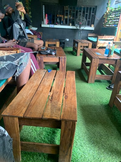 The image shows an outdoor seating area with several wooden tables and chairs placed on artificial turf. There are at least three individuals visible, with one person in the foreground sitting on a wooden bench with their legs crossed, wearing ripped denim shorts and a maroon track pant. Another person, seen from the waist up, is seated at a table, holding a phone and wearing a black jacket. A third person, partially visible on the left, appears to be wearing a patterned headwrap and a black jacket. In the background, a bar area with shelves stocked with bottles and a fan is visible. The lighting suggests it is daytime, with sunlight casting shadows on the ground. The setting appears to be a casual outdoor gathering or a small eatery.