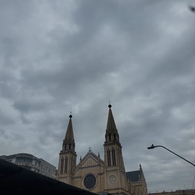 The image depicts the Metropolitan Cathedral Basilica of Curitiba in Curitiba, Brazil, under a cloudy sky. The cathedral's spires and facade are visible, including the clock, rose window, and architectural details. The building is partially obscured by an adjacent building and possibly an overpass or bridge in the foreground. A street light is visible above the cathedral. No people are visible.