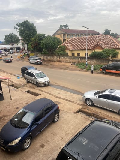 The image depicts a street scene in Jos, Nigeria, under an overcast sky. The foreground features a dark blue Peugeot hatchback parked on a concrete surface. Further down the street, a silver sedan is parked. To the left of the silver sedan, a silver minivan is parked on the side of the road. There are several other vehicles visible on the street, including a black van with orange accents parked further down on the right. A man in a white shirt and dark pants is standing on the roadside, facing away from the camera. The environment includes buildings with corrugated iron roofs, some with yellow facades and others that appear older and weathered. Greenery, including trees and bushes, is present alongside the road and around the buildings. The street surface is paved, with some areas showing signs of wear and puddles. The overall scene suggests an urban setting with everyday activity.