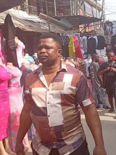 The image depicts an outdoor market scene in Fegge, Nigeria. In the foreground, a dark-complexioned adult male with short dark hair wears a short-sleeved collared shirt patterned with brown, white, and purple geometric shapes. He is positioned slightly off-center to the left, looking towards the viewer's right with a neutral expression. To his left, several clothing items are displayed on mannequins, including a pink form-fitting dress and a multi-colored maxi dress. To his right and in the mid-ground, two other adult males are visible: one in a dark grey t-shirt with light blue jeans, and another in a black t-shirt and shorts, holding a dark object in front of him. The background reveals multiple market stalls or shops with corrugated metal roofs, displaying various garments on hangers and mannequins. Overhead, a dense network of electrical or communication wires spans the scene, above visible multi-story buildings. The ground appears to be an unpaved or roughly paved surface, indicating a commercial street environment.