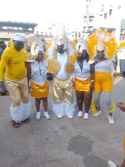 Five people stand in a row, dressed in vibrant carnival costumes, on an asphalt surface in what appears to be a city street. The backdrop includes buildings and a partial view of a stadium.

From left to right: A man wearing a yellow turtleneck shirt, gold pants with white fluffy trim at the bottom, and a white hat. Next to him is a woman in a white top, a yellow skirt, and a large silver and gold headpiece. The person in the middle is wearing a white shirt, gold pants with white trim, and a large gold and white feathered headpiece. To their right, another woman is dressed similarly to the first woman, with a white top, yellow skirt, and a silver and gold headpiece. On the right is a woman wearing a white top, yellow shorts with fringe, gray tights, and a gold and white feathered headpiece. They are all wearing sneakers except the man on the left who wears sandals.

There is a hand holding a smartphone visible on the right, implying someone is taking a picture.

The image is taken in Lagos, Nigeria, as indicated by the architecture and the appearance of the individuals.