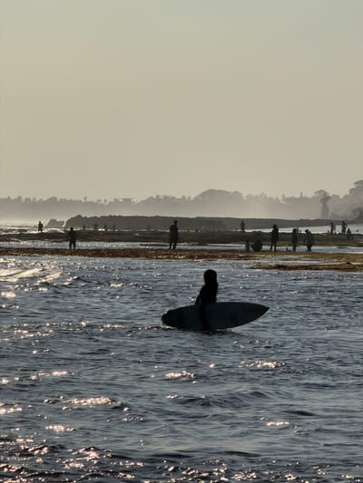 A silhouette of a person sits on a surfboard in the foreground, facing away from the camera, in a body of water. The water has a rippled surface with reflections of sunlight. In the mid-ground, a rocky shoreline is visible, with several silhouetted figures standing on it. Some of these figures appear to be interacting with each other. Further in the background, a hazy coastline with trees and buildings is discernible. The overall atmosphere is serene and suggests a coastal environment. This scene is located in North Kuta, Indonesia.