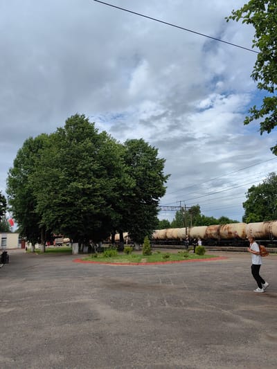 A wide shot captures an outdoor scene at what appears to be a train station in Fanipal, Belarus. The sky is overcast with dramatic clouds, suggesting either early morning or a day with changing weather. In the foreground, a paved area leads towards a grassy island with several trees. Behind this, a long line of train cars, likely tankers, stretches across the frame. Some of these tankers show signs of rust. A few people are visible near the train, and one person, a young male wearing a white t-shirt, black pants, and white sneakers, is walking across the paved area in the right foreground, carrying a brown backpack. His posture suggests movement, possibly heading somewhere. To the left of the frame, a small building with a red-trimmed roof and a flag is visible. The overall atmosphere is somewhat muted due to the cloudy weather, but the presence of people and the train suggests a functional, active location. One tanker car has the text "748998127" visible.