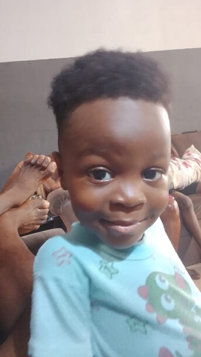 A young child with dark skin, close-cropped dark hair, and wearing a light blue shirt with a cartoon design is the central subject of the image. The child is smiling slightly and looking directly at the camera. In the background, out of focus, are human feet and legs resting on what appears to be a brown upholstered couch. The wall behind is two-toned, with grey on the bottom and off-white on top. Given the request for a location of Lokoja, Nigeria, this scene likely represents a casual domestic setting there.