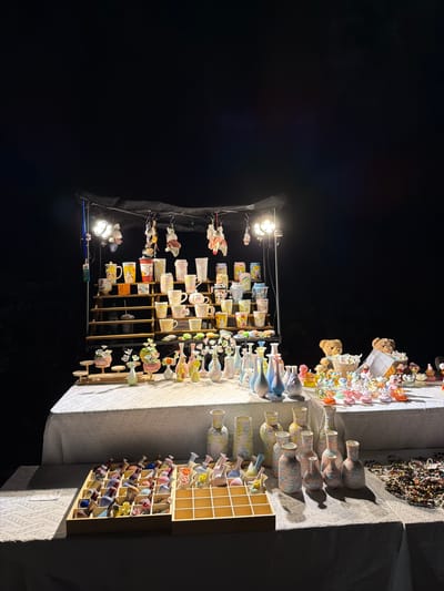 This image captures a nighttime scene at a stall in Chongqing High-tech Zone, China, featuring a variety of handcrafted items. The stall is illuminated by spotlights, casting a warm glow on the merchandise. Displayed on tiered shelves and tables are numerous ceramic mugs adorned with cartoon characters, small decorative vases in pastel colors, and various miniature figurines. Two teddy bear figures sit on a table in the background, surrounded by small decorative items. The stall is covered by a dark canopy, and the surrounding area is dark, suggesting an outdoor market or event. The items suggest a focus on decorative ceramics and collectibles, likely for sale.