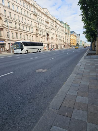 The image shows a street in Moscow, Russia, on a cloudy day. The street is wide and paved with asphalt, with white lane markings. On the left side of the street, there are several large, multi-story buildings with ornate architecture and many windows. A white tour bus is parked on the side of the road. On the right side of the street, there is a sidewalk made of stone pavers, lined with trees. Several yellow taxis are visible on the street, moving away from the camera. A few pedestrians are walking on the sidewalk. The overall scene depicts a typical urban streetscape in Moscow.