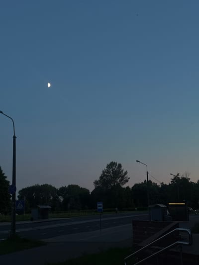 The image depicts a street scene in Minsk, Belarus, during twilight. The sky is a deep blue, with a sliver of the moon visible in the upper left quadrant. A street lamp stands prominently on the left side of the frame, its dark pole and curved arm silhouetted against the sky. Across the street, there are trees and a bus stop shelter. Further down the road, another street lamp is visible. The road itself is marked with white lines. A pedestrian crossing is present in the foreground. A brick structure with a metal railing is in the lower right corner, suggesting an entrance or stairway. A few figures are faintly visible in the distance, walking along the sidewalk. The overall atmosphere is calm and serene, characteristic of the evening hours in an urban setting.