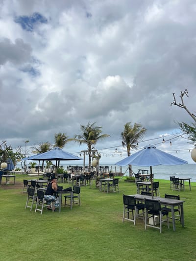 This media file depicts an outdoor seating area of a restaurant or cafe, likely near a beach, given the presence of palm trees and the ocean in the background. The scene is set on a well-maintained grassy lawn with multiple wooden tables and chairs arranged in rows. Several large, light blue umbrellas are set up to provide shade over some of the seating areas. String lights are visible above, suggesting the space is also used in the evening.

One individual is seated at a table, leaning back in a chair, wearing a black tank top and patterned shorts. Their posture suggests relaxation.

The sky is heavily overcast with dramatic, dark clouds, indicating a potentially stormy or moody weather condition. Despite the clouds, there is ambient light, suggesting it is daytime. The location cues, such as the palm trees and the ocean, along with the provided context of Mengwi, Indonesia, point to a tropical or subtropical coastal setting. The overall atmosphere appears to be one of quiet leisure, possibly before or after a meal, amidst a dramatic sky. No visible text is present in the image.