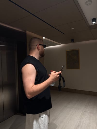 In a dimly lit indoor setting, possibly a hotel hallway or elevator, a person is seen from a low-angle, rotated perspective. The subject is a bald man with a beard, wearing a black sleeveless shirt and grey sweatpants. He is holding a smartphone in his right hand and a water bottle in his left. Sunglasses are perched on his head. The background features wood-paneled walls and a tiled section, suggesting a modern architectural style. A framed picture hangs on one of the walls. The lighting is subtle, with some light fixtures visible on the ceiling and walls. The overall mood appears casual and relaxed, as the man is engrossed in his phone. There are no clear indicators of the time of day or weather, but the indoor environment suggests it could be any time. The location context of Dubai, United Arab Emirates, doesn't offer specific visual cues in this particular image.