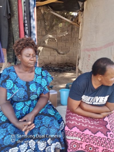 The image captures two adult women seated outdoors in Nairobi, Kenya, within what appears to be an informal, possibly market, setting. The photo is rotated, showing the subjects from a slightly elevated perspective.

In the foreground, a woman with short, curly brown hair is seated, looking downwards with a calm or pensive expression. She wears a striking blue and black patterned dress, and her hands are clasped in her lap. To her immediate left, a stack of various colored fabrics or clothing items, including red, green, and black pieces, is visible.

Behind and to the right of the first woman, another woman is seated. She is seen from the back and side, wearing a dark t-shirt with partially visible white text that reads "FORTH AVE". Her arms are crossed, and a vibrant red and pink patterned fabric is draped over her lap. Her head is turned away from the camera. A light blue plastic bucket is positioned behind her, next to a light-colored rectangular container.

The background reveals a makeshift shelter structure with a ceiling made of a white tarp or sack material, on which the text "MIXED B" is partially discernible. Wooden beams support this overhead covering. Beyond the immediate structure, natural elements like tree branches and a bare trunk are visible against a textured wall constructed from rectangular blocks or bricks. The lighting indicates it is daytime with clear weather. Both women appear to be resting or waiting.

A digital watermark in the bottom right corner reads "Samsung Dual Camera" and "Shot with my Galaxy A03".