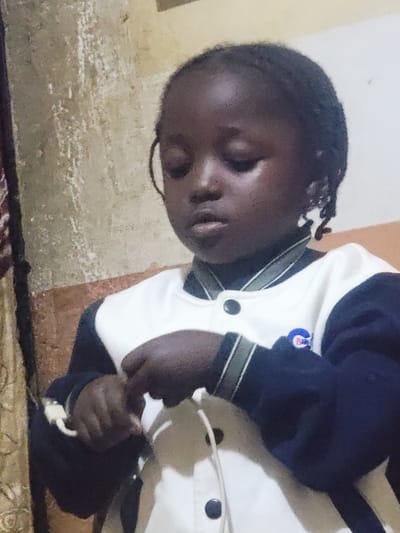 A young child with dark skin and braided hair is the subject of this portrait. The child is wearing a navy blue and white jacket with green trim. The child's hands are held together in front of their chest, and they appear to be holding a white string. The background of the image is a textured wall that is a mix of light brown and white. The lighting is dim, casting shadows on the child's face. The image appears to have been taken indoors, possibly in a home environment within the city of Barakin Akawo, Nigeria.