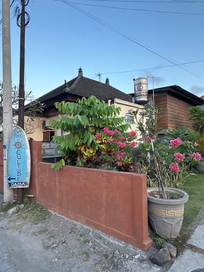 In North Kuta, Indonesia, a light blue surfboard-shaped sign is mounted on a utility pole. The sign features a floral design at the top, followed by the word "THE" in capital letters, and then "CULTURE" written vertically. Below "CULTURE" is an arrow pointing to the left, and the word "DAMA". Behind the sign, a red-brown wall encloses a garden with vibrant pink bougainvillea flowers and lush green foliage. A house with a dark roof and beige walls is visible behind the garden, with a large water tank on its roof. A wooden-paneled section of the house is to the right. The ground in front of the wall is covered in gravel and dirt. A large, grey planter with a gold Greek key pattern sits to the right of the wall. Electrical wires are strung across the clear blue sky above.