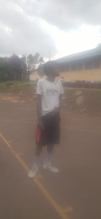 A person stands on a paved area with a yellow line. They wear a white shirt with "ENIGMA" printed on it, black shorts, and white shoes. The background shows a cloudy sky and some greenery. The photo appears slightly blurry and is rotated counter-clockwise. The overall lighting seems overcast.