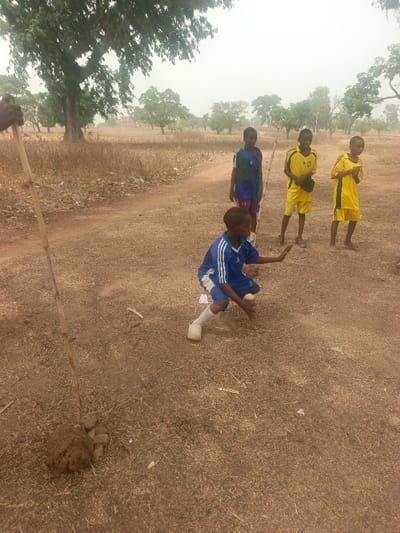 Four children stand in a dry, grassy field. One child in blue and white kneels, holding a white object, possibly a ball. Three other children stand behind him; one wears blue and the other two wear yellow soccer uniforms. In the foreground, a slender tree branch leans against a pile of earth. Trees dot the landscape in the background under a pale sky. A dirt path runs through the field.