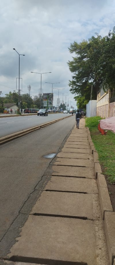 The image shows a street in Kisumu, Kenya, at coordinates -0.103 latitude and 34.759 longitude.  A man walks on a concrete sidewalk on the right side of the image. There is a small puddle on the asphalt road in the middle of the frame. The road is lined with street lamps and there are a few cars in the distance. There are some trees on the right side of the image and a brick wall with a red tarp in the foreground.  There is green grass along the sidewalk.  There is a large billboard on the left side of the image and a small, single-story building.  The image is taken from the perspective of a person standing on the sidewalk looking down the street.