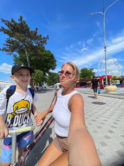 In Anapa, Russia, a woman and a boy stand on a paved promenade under a bright blue sky. The woman, with blonde hair and sunglasses, wears a white tank top and brown shorts. The boy, wearing a black cap, a white t-shirt with a cartoon duck, and blue shorts, holds a scooter. They are holding hands, and the woman is taking a selfie. In the background, a Ferris wheel and carnival rides are visible, along with trees and other people walking. The scene appears to be a sunny day in a seaside resort.