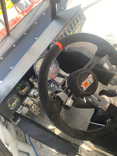 This is a close-up interior shot of a race car cockpit. The steering wheel, covered in a black, suede-like material with a red accent at the top, dominates the foreground. The steering wheel features a center hub with the MPI logo. Behind the steering wheel, the car's seat and safety harness are visible.

Various gauges and switches are mounted on a panel to the left of the steering wheel. The gauges display information such as oil pressure, water temperature, and engine RPM. The switches include labels like "FUEL". The pedals are also visible near the bottom of the frame.

The car's interior is made of metal, with a diamond-plate pattern on the floor. Blue and black wires are visible within the car. A Telex PT-300 device is mounted nearby the steering wheel.

The image does not offer any information to allow determining the location in the city of San Bernardino Tlaxcalancingo, Mexico.