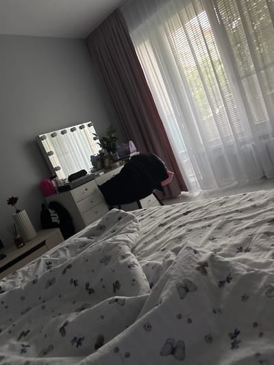 The image displays an indoor scene, likely a bedroom or living space, captured from an elevated perspective and presented in a rotated orientation. The setting is a bright room, indicative of daytime, with sunlight filtering through a large window covered by sheer white blinds. Beyond the blinds, blurry green foliage suggests the presence of trees or plants outside. Below the window, a darker, possibly pinkish-brown, curtain is partially visible.

A prominent feature is a bed covered with rumpled white bedding, adorned with a pattern of butterflies and small dots or floral motifs in shades of grey and light blue. To the left of the bed, against a light grey wall, stands a white dresser or vanity. On its surface, various objects are present, including a black item (possibly clothing or a bag), a pink container, and a small green potted plant. A rectangular Hollywood-style vanity mirror with light bulbs around its frame leans against the wall, reflecting the window blinds. Faint text, "OFFICE STYLE," is visible on an item on the dresser.

On the floor beneath the dresser, a black bag with the partially visible text "JUST DO IT" is placed. Further into the room, another piece of white furniture, possibly a side table, holds a lamp with a ribbed white shade and a small container with diffuser sticks. The ceiling, particularly in the top-left corner of the image, appears to have a dark, possibly wood-paneled finish. The overall scene suggests a lived-in and peaceful environment in Sofia, Bulgaria, with no specific activity taking place.