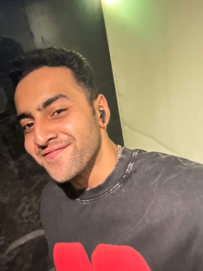 A man is shown in a selfie with a dark, textured background and a lighter wall on the right. He is wearing a dark gray, distressed sweatshirt with a large red graphic on the chest, a silver chain necklace, and black earbuds. He has dark hair, dark eyes, and a light beard. He is smiling and looking at the camera. The lighting suggests an indoor setting, possibly in a residential or casual public space in Kolkata, India. The overall mood is relaxed and informal.