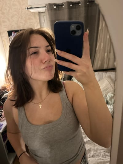 A young woman takes a selfie in a mirror. She has brown hair, fair skin, and wears a grey tank top. A gold cross necklace hangs around her neck. She holds a dark blue iPhone in her left hand, her long nails visible. The background reveals a bedroom setting with curtains, bedding, and a glimpse of furniture. The image is slightly rotated.