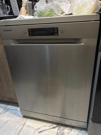 This image shows a stainless steel Samsung dishwasher. The top control panel has several buttons and icons, including a power button, a clock icon, a half-full icon, a lock icon, and a "BAŞLAT" button which appears to mean "START" in Turkish. Below the control panel is the main door of the dishwasher. The dishwasher is situated in a kitchen environment with light-colored tiled flooring and wooden cabinetry to the left. On top of the dishwasher, there are various kitchen items, including a glass pitcher, a plastic bag with some sort of food item inside, and another plastic bag containing what appears to be produce. The scene is indoors. The image does not provide any information that would allow for the determination of its location in Tbilisi, Georgia.
