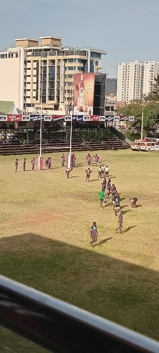 A rugby match on a grassy field is captured from an elevated perspective. Teams in maroon and black uniforms are visible. Spectator stands line the field's edge, adorned with banners and advertisements. Beyond the stands, buildings of varying heights, including a prominent billboard, mark the cityscape. The sky is clear, indicating daylight. Part of a structure or railing is visible in the lower right foreground, adding a sense of depth to the scene.