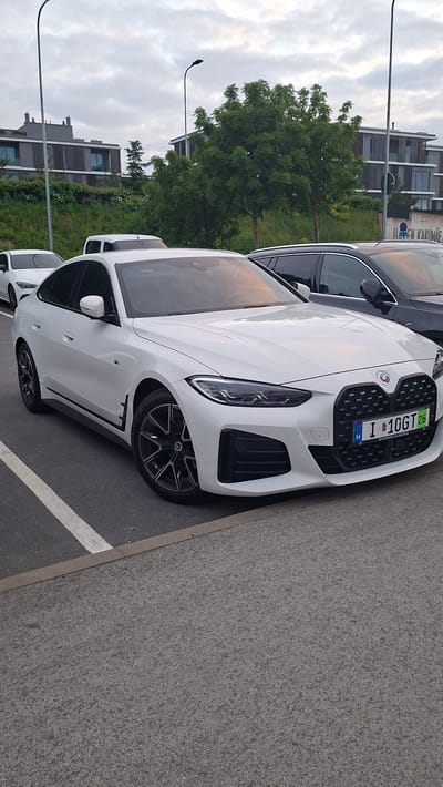 A white BMW i4 electric vehicle is parked in a lot in Tallinn, Estonia. The car is the primary subject, positioned diagonally in the frame with its front end facing the camera. The vehicle is clean and appears to be in good condition. The license plate is visible and reads "I 10GT26". In the background, several modern buildings with large windows and balconies are visible, partially obscured by green trees. The sky is overcast with a mix of gray and white clouds. Another dark-colored car is parked to the right of the BMW. The parking lot surface is asphalt, marked with white lines indicating parking spots. Streetlights are also present in the scene.