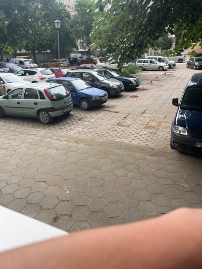 The image shows a street with cars parked on the side. The street is paved with hexagonal concrete blocks, and there are trees and buildings in the background. The cars are of various colors and models, and they are parked in a line. Some of the cars appear to be old, while others are newer. There is also a lamppost in the image. The image is taken from a high angle, looking down at the street. It appears to be daytime, and the weather is overcast. The scene is a typical urban street in Plovdiv, Bulgaria, with residential buildings and greenery. No people are clearly visible. There are no notable emotional cues. Some visible text on a license plate appears to read "PB".